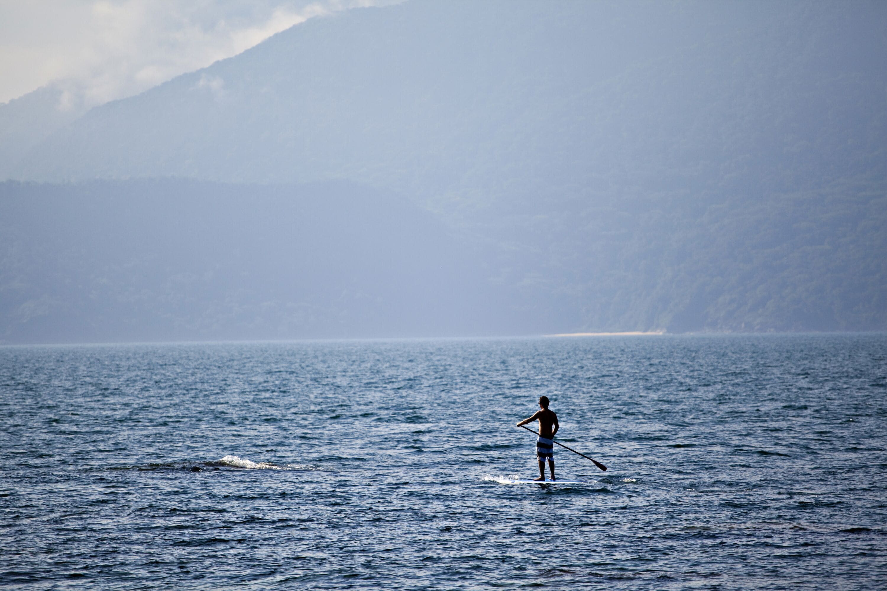 A person paddleboarding on a calm sea with mountains in the background.