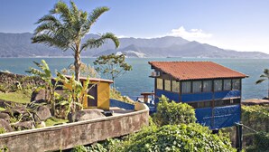 A coastal building with a red roof overlooking a body of water and mountains. - Pousada Isola Bella - Sobre o Mar (Ilhabela)