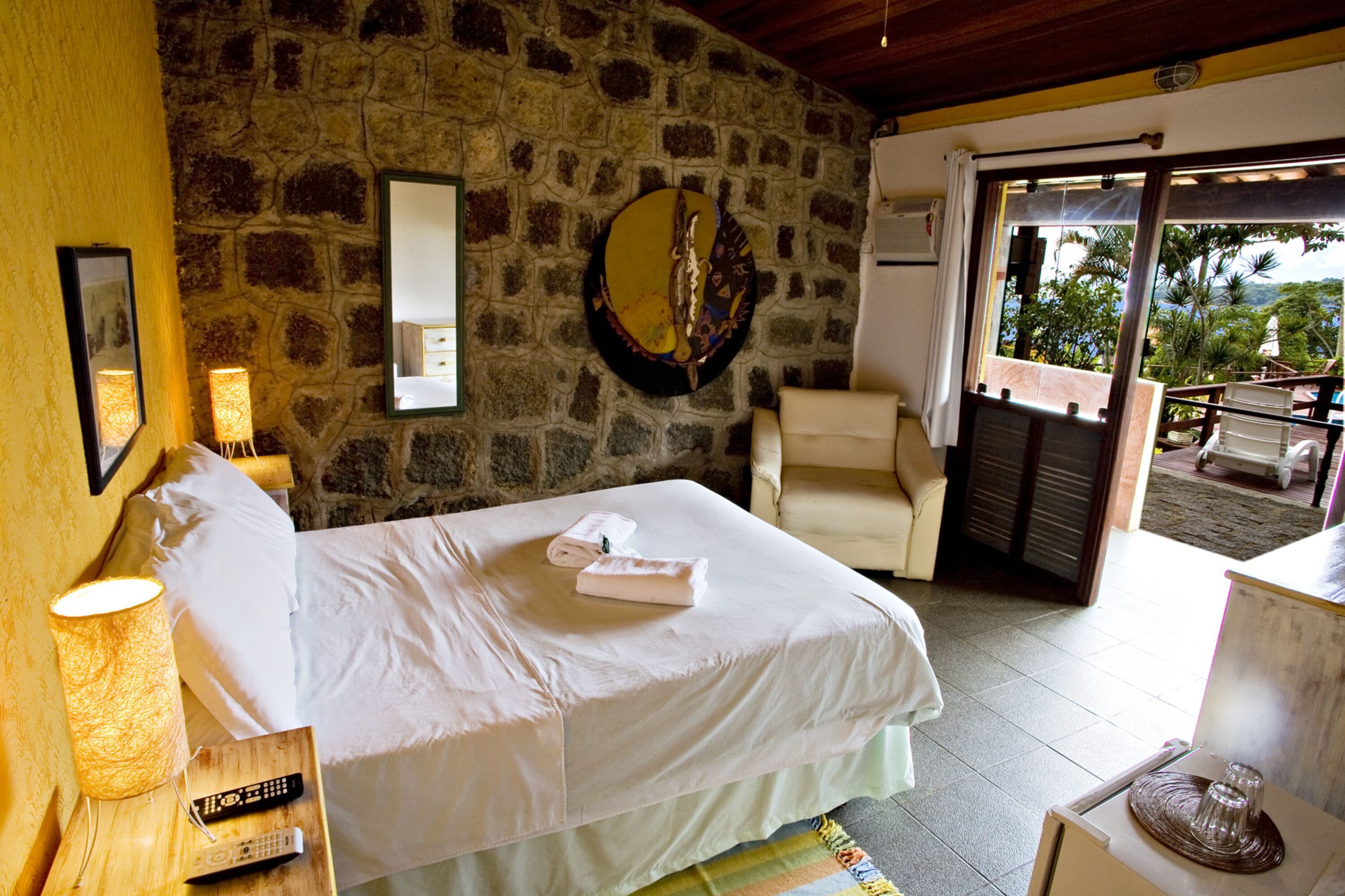 A bedroom with a stone wall, a bed with white linens, a bedside lamp, a chair, and a view through a glass door.