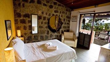Une chambre avec un mur en pierre, un lit recouvert de draps blancs, une lampe de chevet, une chaise et une vue sur l’extérieur, visible à travers une porte vitrée.