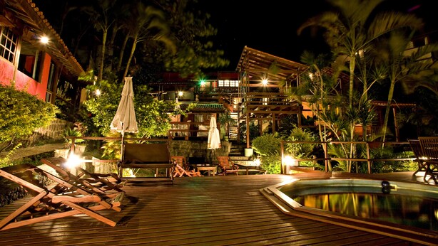 Eine Holzterrasse mit Liegestühlen und einem Pool, umgeben von tropischen Pflanzen und einem mehrstöckigen Gebäude mit beleuchteten Fenstern.