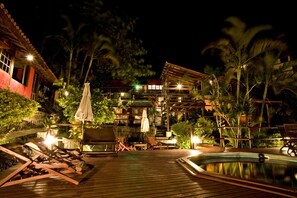 Terraza de madera con sillones de descanso y piscina, rodeada de plantas tropicales y un edificio de varios pisos con ventanas iluminadas.