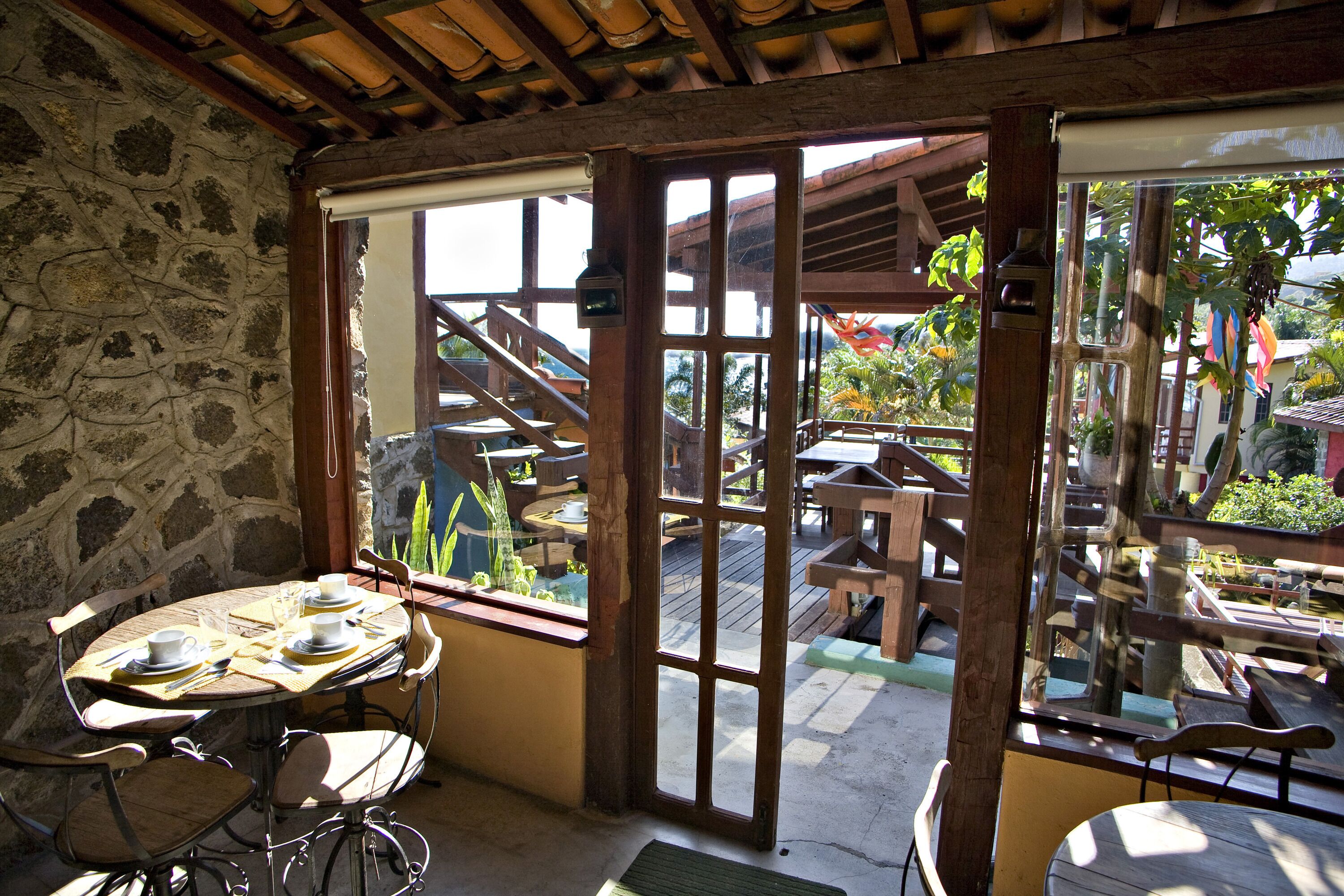 A stone-walled room with a wooden table set for two, a window with a view of outdoor wooden furniture and a staircase leading to a deck.