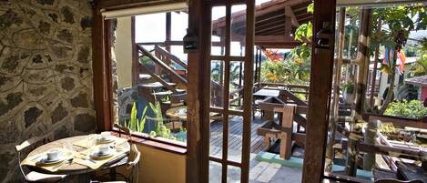 A stone-walled room with a wooden table set for two, a window with a view of outdoor wooden furniture and a staircase leading to a deck.