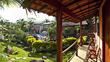 Guests can enjoy the serene outdoor space with a wooden deck, chairs, and a table amidst lush greenery and a traditional building in the background.