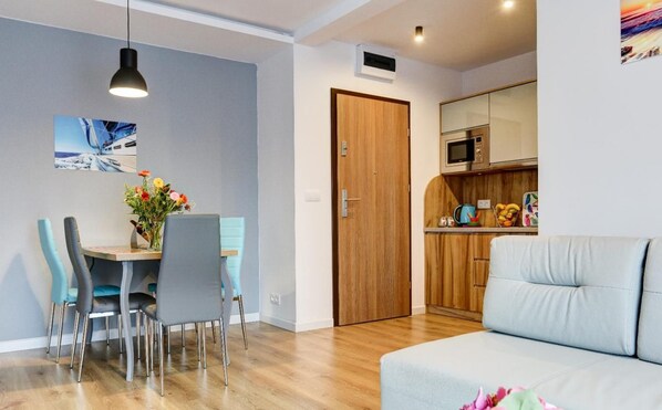 A modern living room with a sofa, a dining area with chairs and a table, a wooden door, and a kitchen area with a microwave.