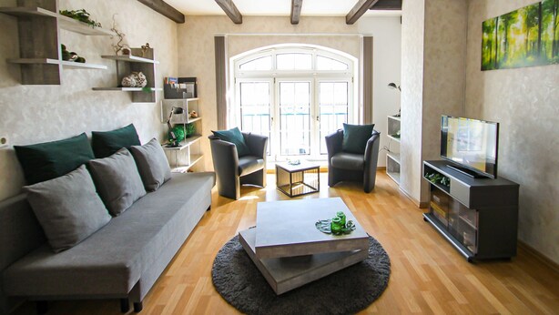 A modern living room with a grey sofa, a coffee table, and a TV stand. There are two armchairs and a bookshelf. The room has wooden flooring and a large window with a view of the outside. - Apartments Vogelsang - 3-room apartment (Kramerhof)