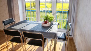 A dining area with a table and chairs, a potted plant, and a view of a yellow field through large windows. - Apartments Vogelsang - 3-room apartment (Kramerhof)