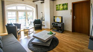 A spacious living room with a large window, a fireplace, and a television. - Apartments Vogelsang - 3-room apartment (Kramerhof)