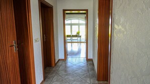 A hallway with wooden doors and tiled flooring leading to a room with a large window and a chair. - Apartments Vogelsang - 3-room apartment (Kramerhof)