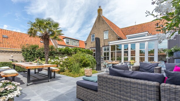 An outdoor patio area with a wicker sofa, a wooden table, and a dining set. - De Herberg (Renesse)