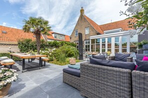 An outdoor patio area with a wicker sofa, a wooden table, and a dining set. - De Herberg (Renesse)