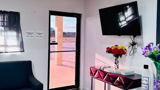 Habitación de hotel con televisor de pantalla plana montado en la pared, una pequeña mesa con un florero, un sofá negro y un ventanal con vistas a un paisaje desértico.