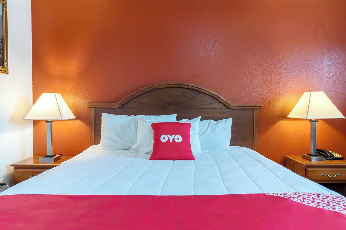 Une chambre d’hôtel avec un lit, deux lampes de chevet, une tête de lit en bois et un oreiller rouge orné du logo « OYO ».