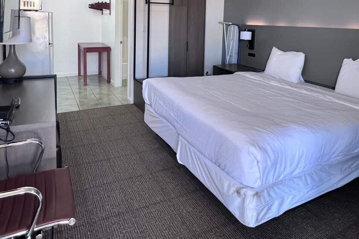 Une chambre d’hôtel avec un grand lit, un bureau avec une chaise, une armoire et une table de chevet avec une lampe.