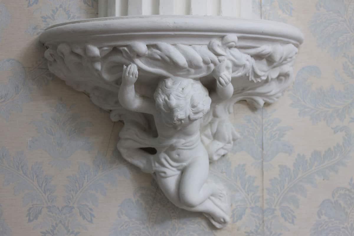 En vit arkitektonisk detalj med en utskuren putto, placerad mot en bakgrund av mönstrad tapet.