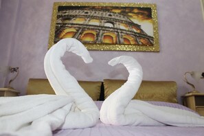 A hotel room with a bed, two white towels arranged in swan shapes, a gold-framed picture of the Colosseum, and a purple wall. - B&B San Peter's Golden Rooms (Rome)