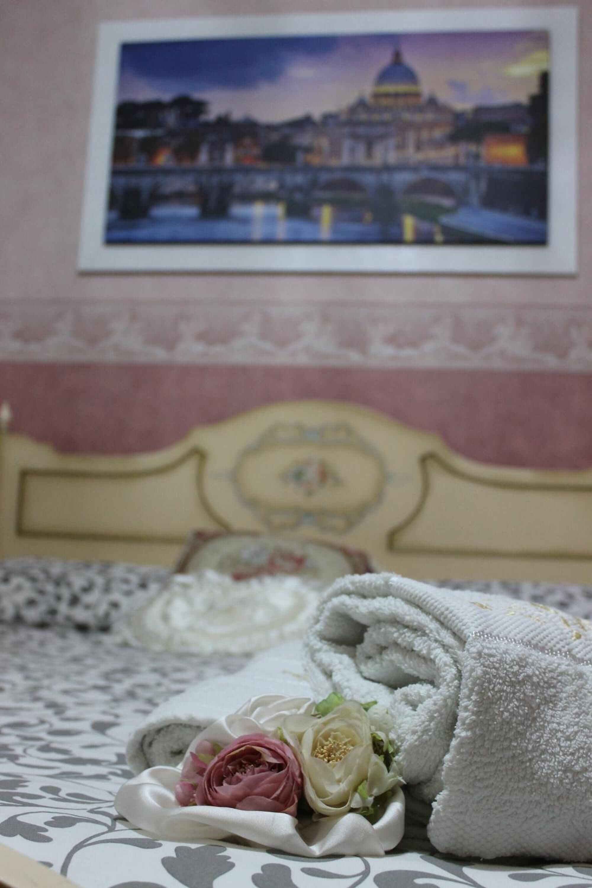 A neatly made bed with a folded towel and a bouquet of flowers, a framed picture of a cityscape on the wall, and a patterned bedspread.