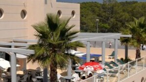 An outdoor seating area with palm trees and umbrellas, a building with circular windows, and a pedestrian walkway. - Les Parasols d'Argens (Roquebrune-sur-Argens)