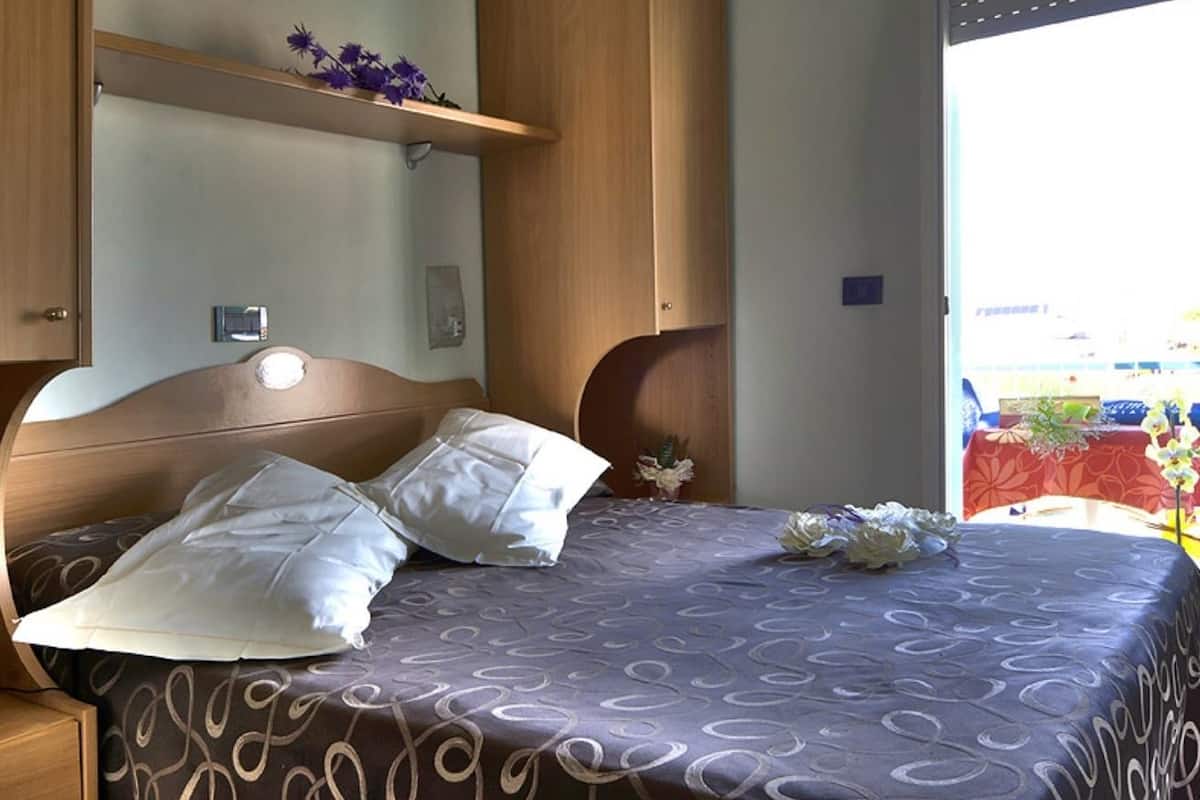 A neatly made bed with a patterned bedspread and two pillows, a wooden headboard, a small bedside table, and a shelf with flowers.