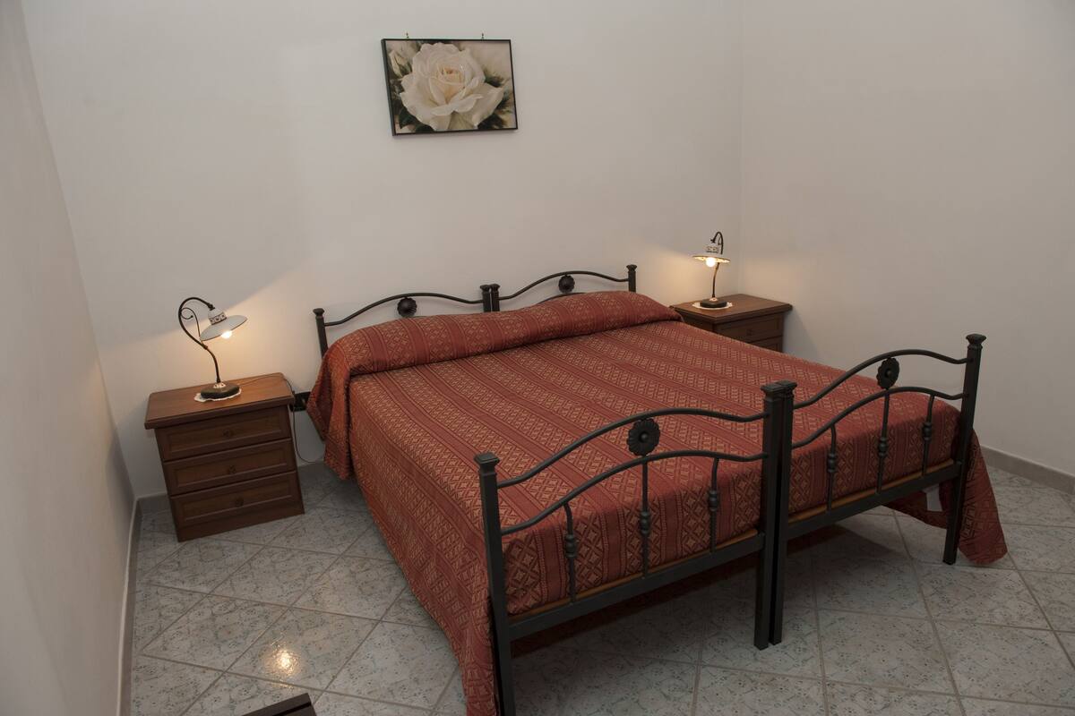 Ein Einzelbett mit gemustertem Bettbezug, zwei Nachttische mit Lampen, eine kleine Holzkommode und ein gerahmtes Bild an der Wand.