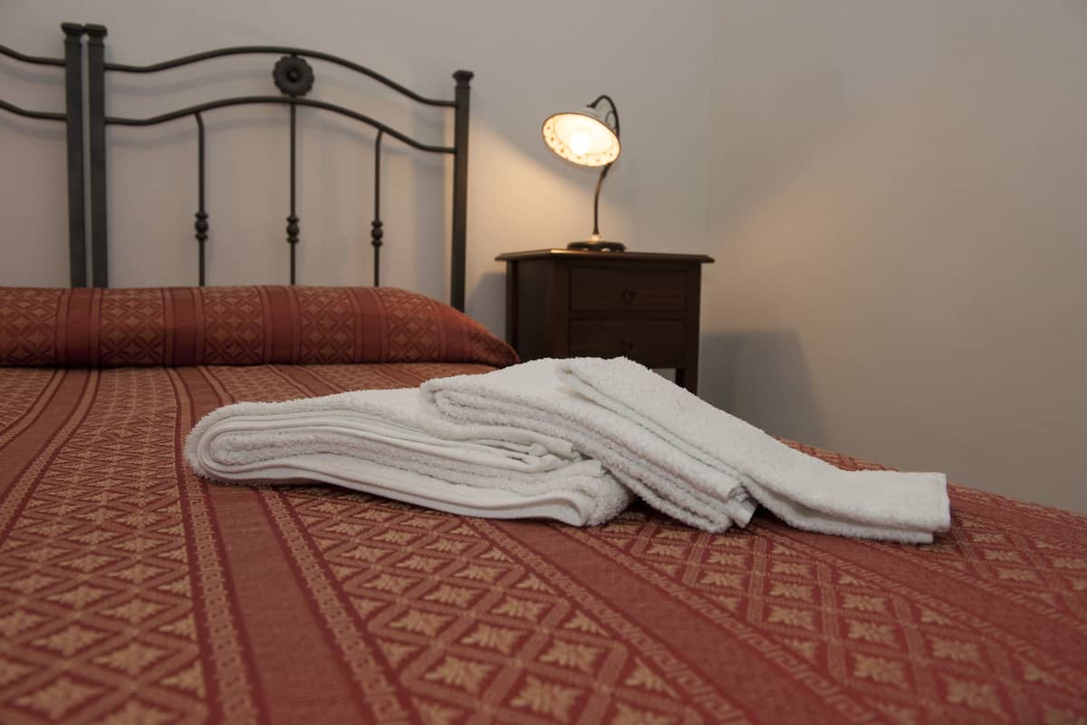 Ein ordentlich bezogenes Bett mit einer gemusterten Tagesdecke, ein gefaltetes weißes Handtuch und ein Nachttisch mit einer Lampe.