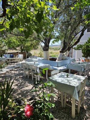 An outdoor seating area with tables and chairs, a large tree, and a view of the landscape. - Alize Otel (Bozcaada)