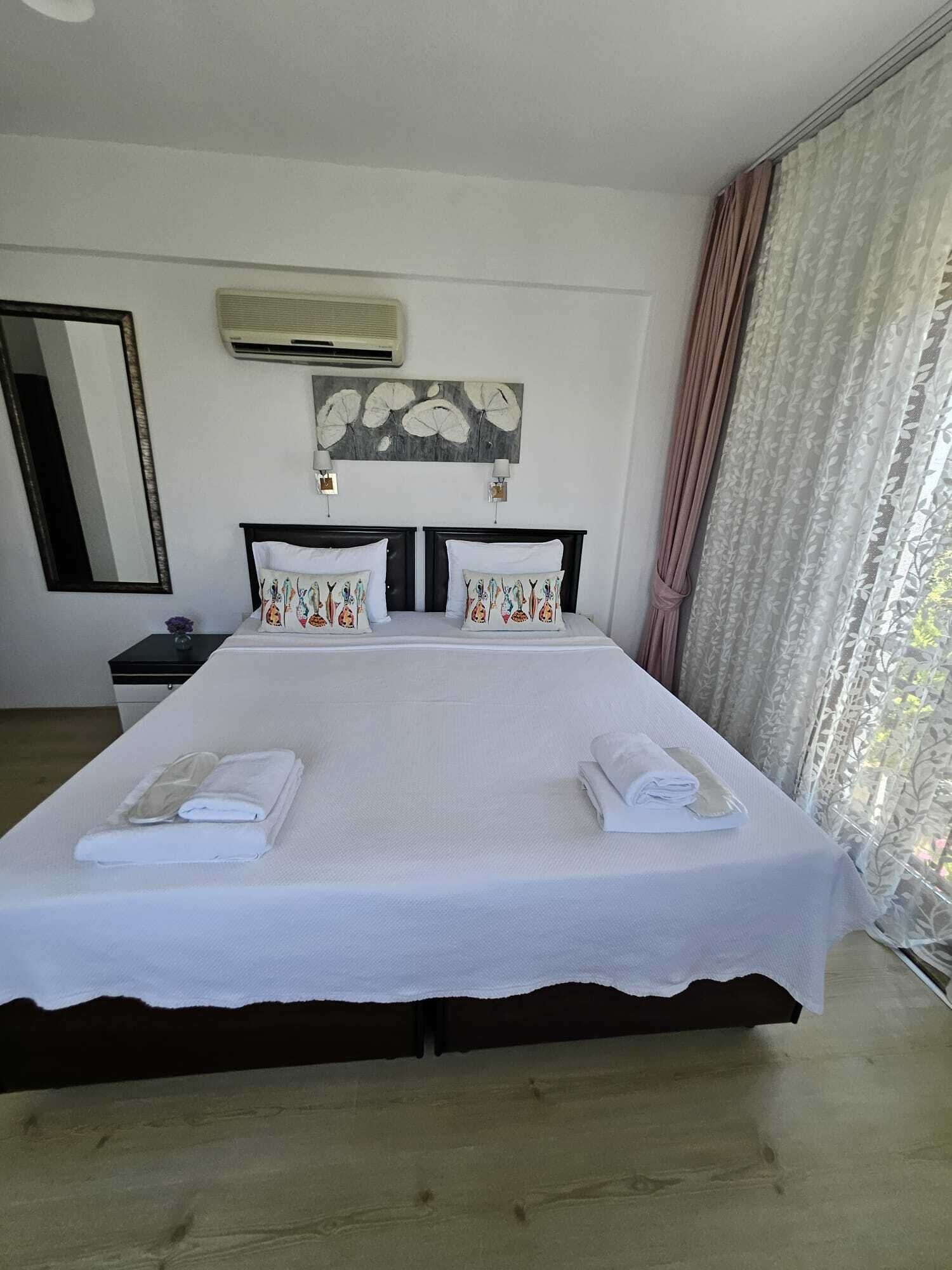 Una cama grande con ropa de cama blanca y almohadones decorativos, una mesita de noche, un espejo y un aire acondicionado de pared.