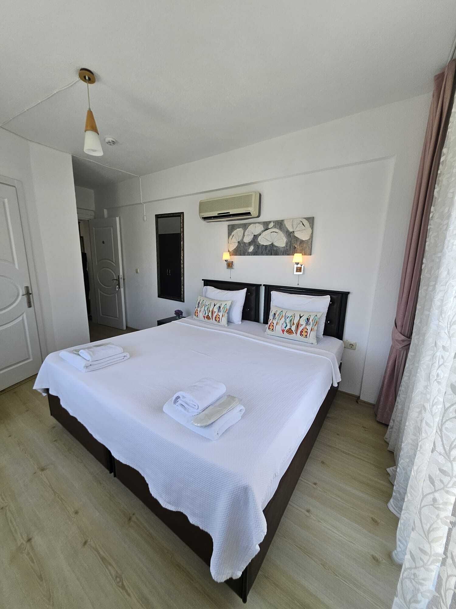 A double bed with white linens, a bedside table, a wall-mounted air conditioner, and a painting above the bed.