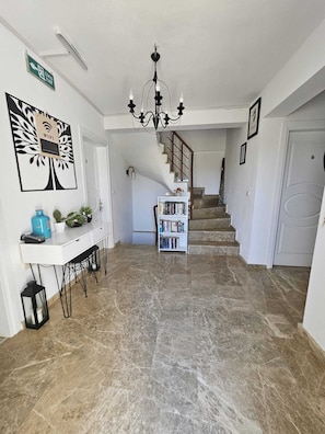 A spacious hallway with a marble floor, a staircase, a desk with a plant, a bookshelf, and a chandelier. - Alize Otel (Bozcaada)