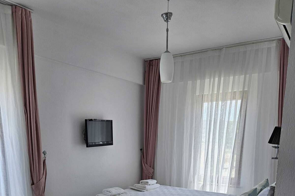 Un dormitorio con cama, televisor montado en la pared, mesita de noche con lámpara y cortinas de gasa.