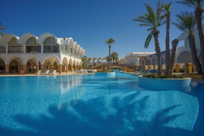 A large swimming pool with sun loungers and palm trees, surrounded by a white building with arched windows.