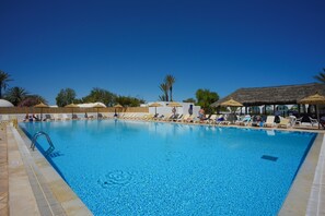 Una piscina azul cristalina con sillas de descanso y sombrillas, rodeada de palmeras y un cielo despejado.