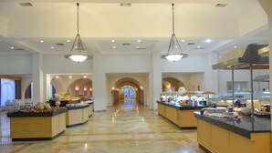 Un amplio vestíbulo de hotel con un pasaje central arqueado, varias estaciones de buffet y una lámpara de araña moderna.