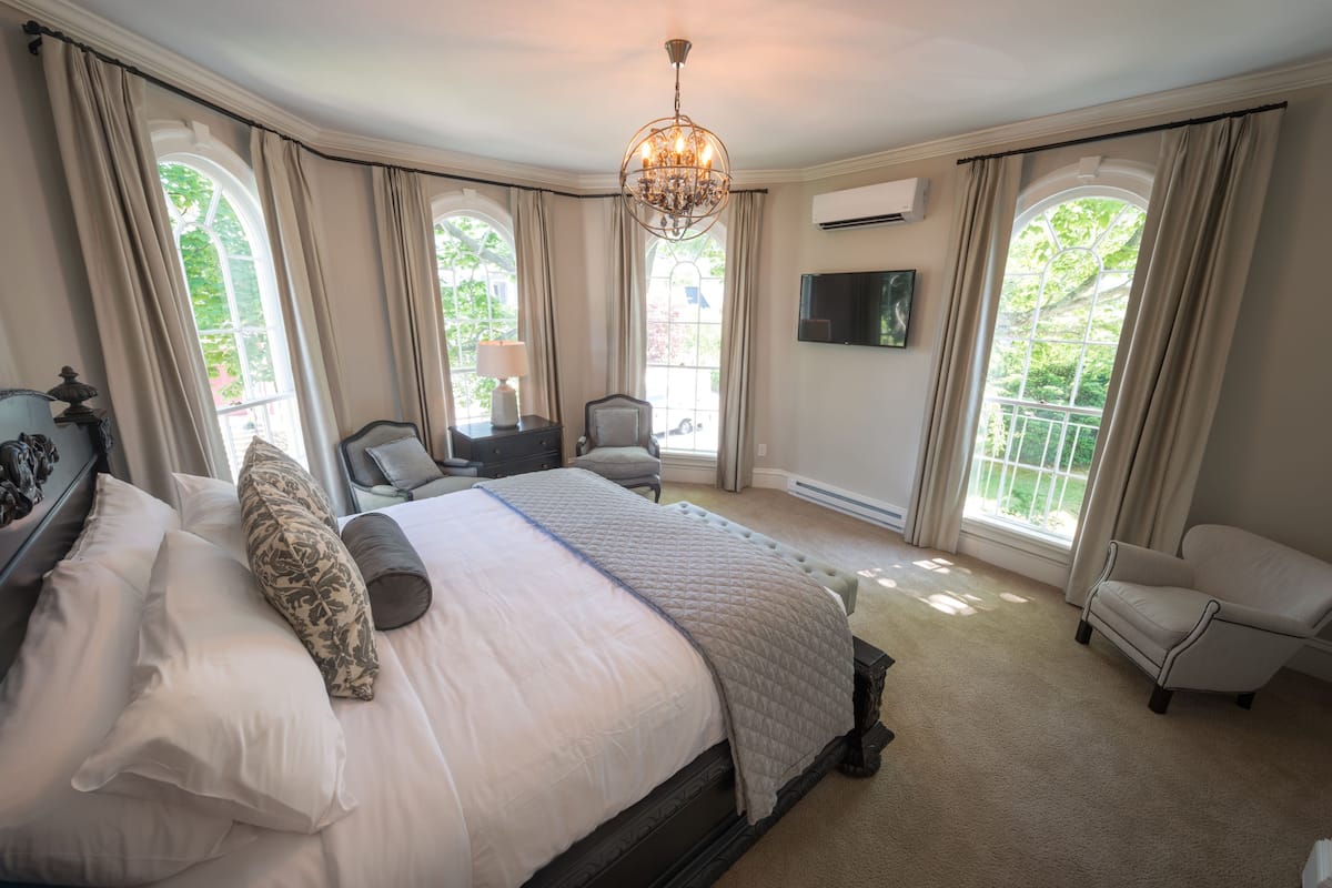 A spacious bedroom with a large bed, a chandelier, a TV, and a view of greenery through the windows.