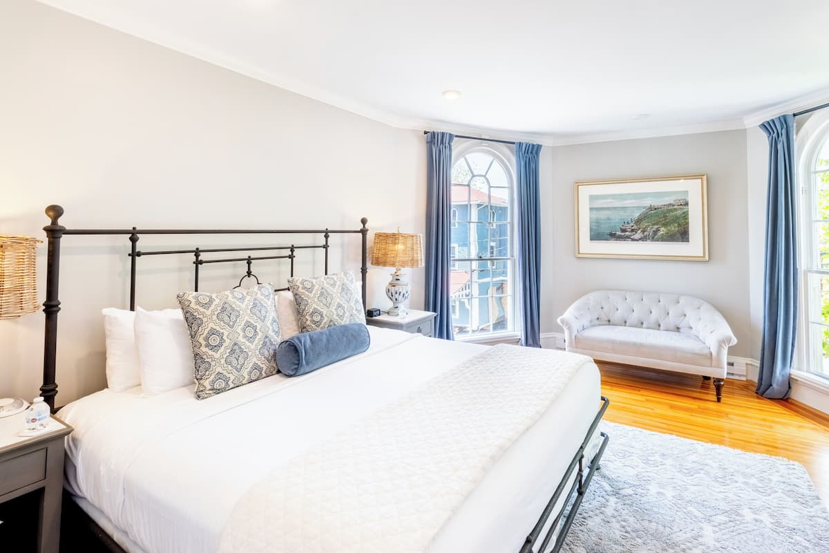 A neatly arranged bedroom with a large bed, a bedside table, a lamp, a chair, and a window with curtains.