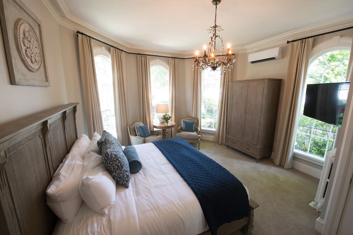 A bedroom with a large bed, a chandelier, a TV, and a view of greenery outside.