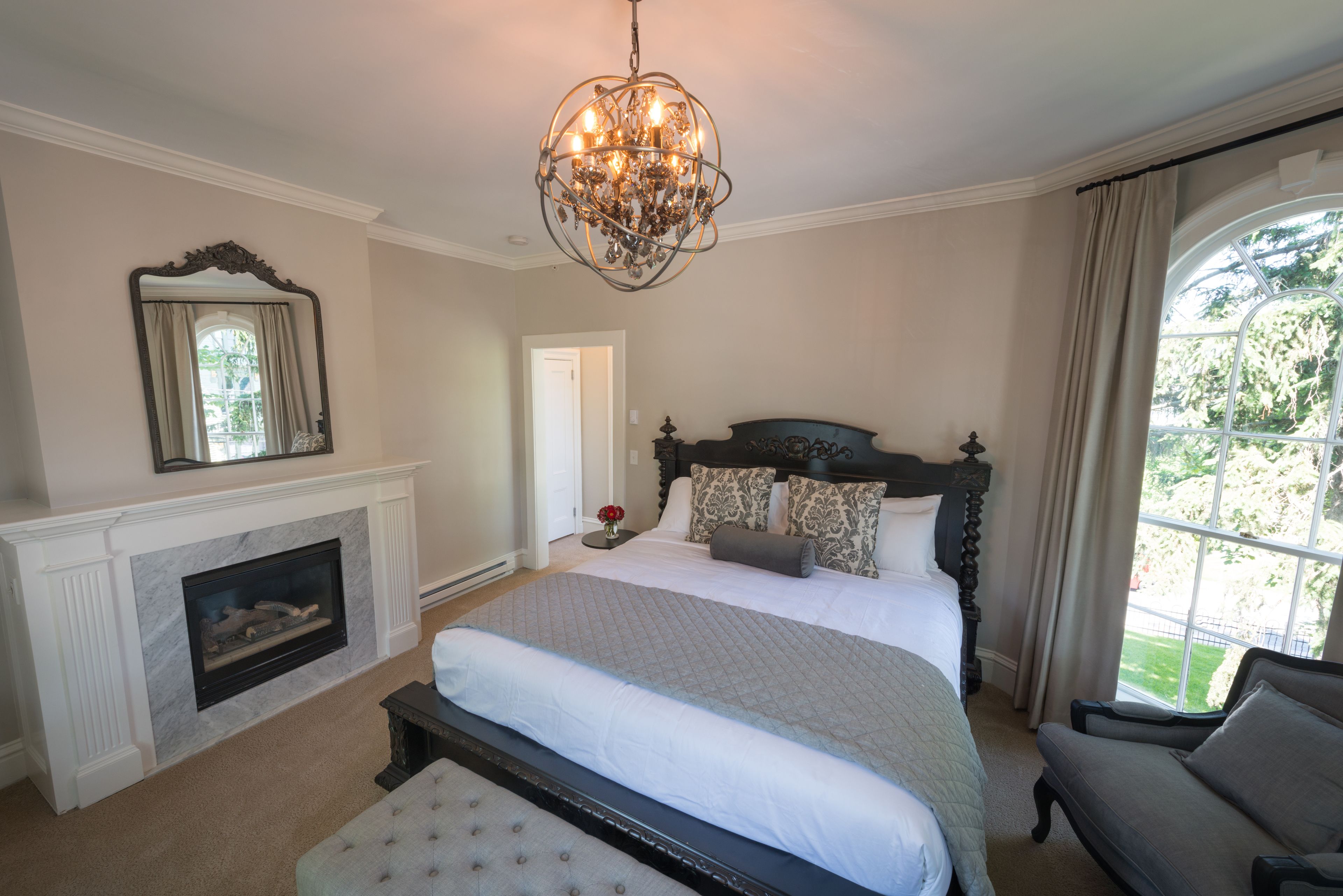 A bedroom with a large bed, a fireplace, a chandelier, and a view of the outdoors.