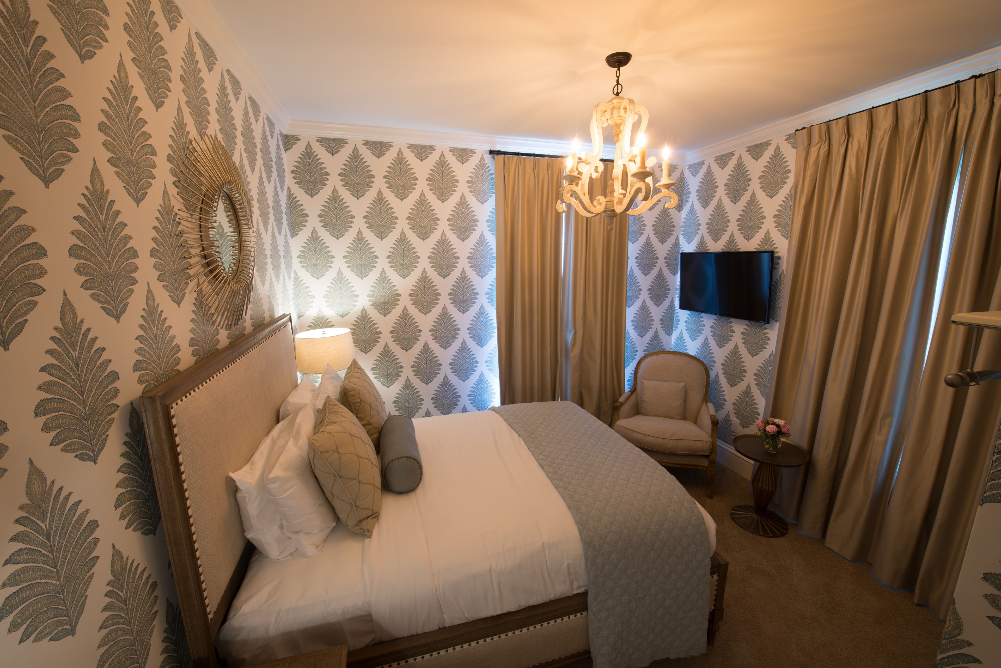 A neatly made bed with pillows, a bedside lamp, a chair, a small table with flowers, and a TV on the wall.