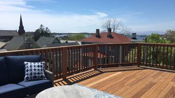 A wooden deck with a round table and blue sofa, overlooking a town with buildings and a church steeple.