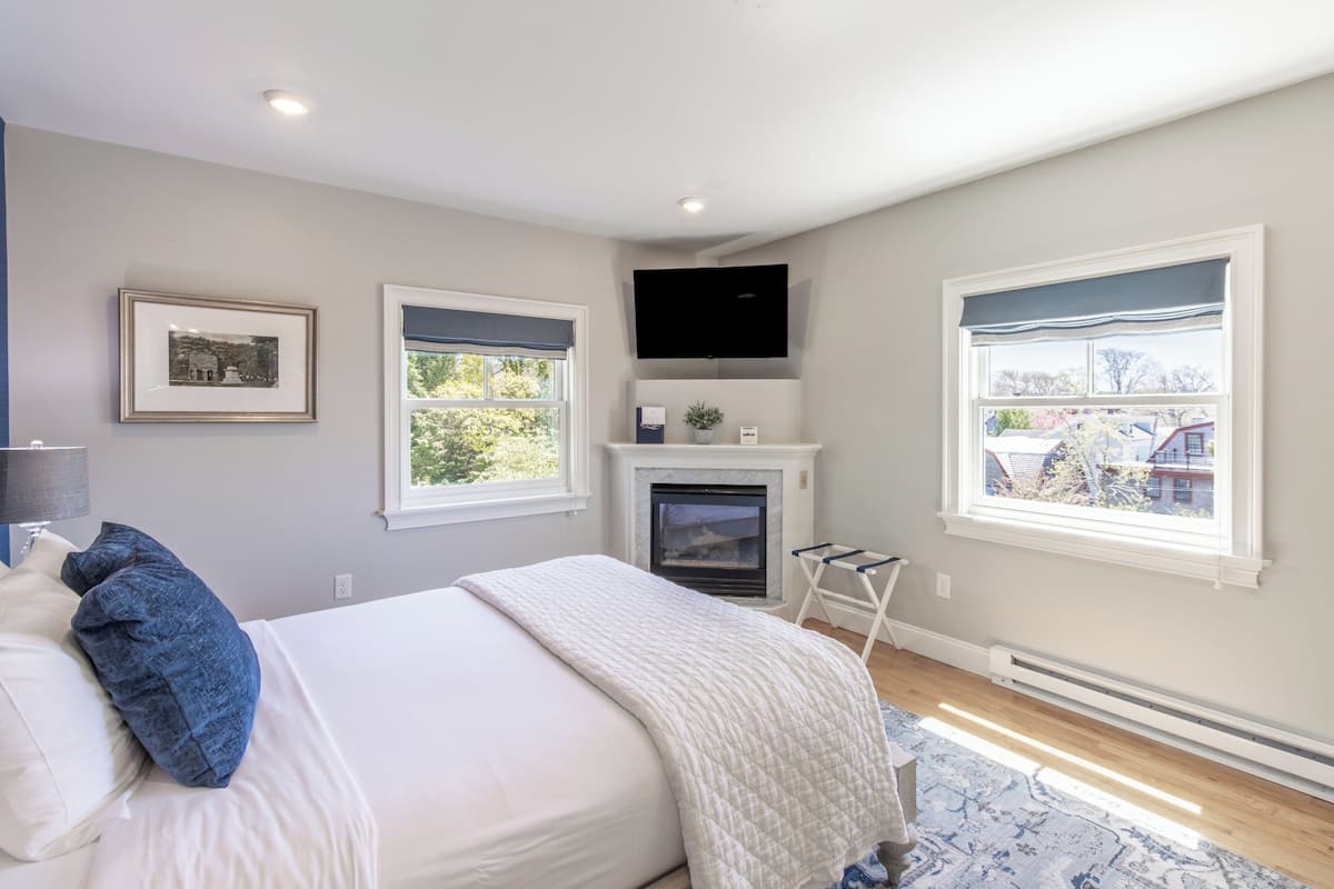 A bedroom with a bed, a fireplace, a mounted TV, and windows with blinds.