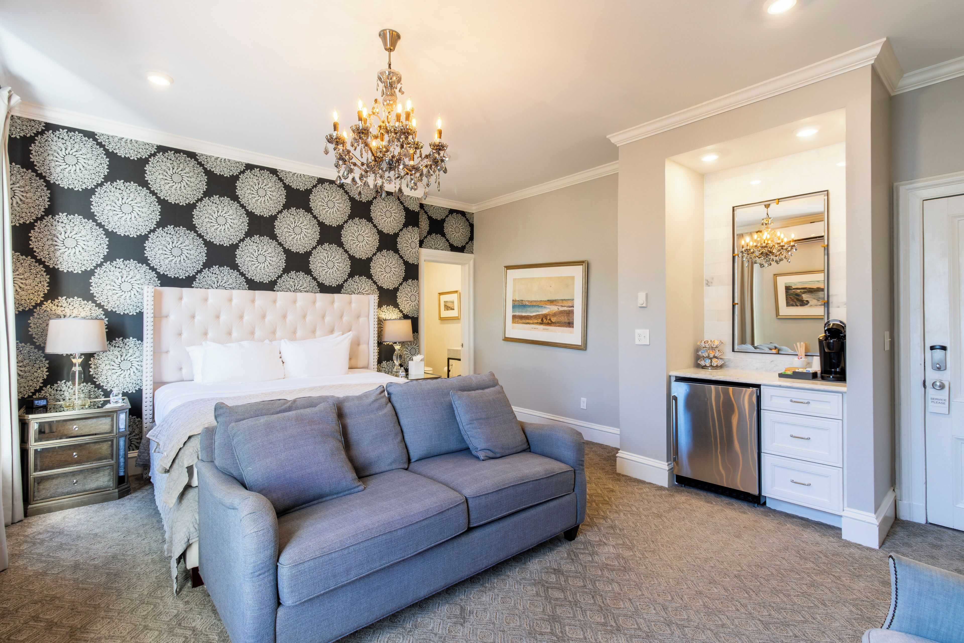 A bedroom with a tufted headboard, a grey sofa, a chandelier, a minibar, and a large mirror.