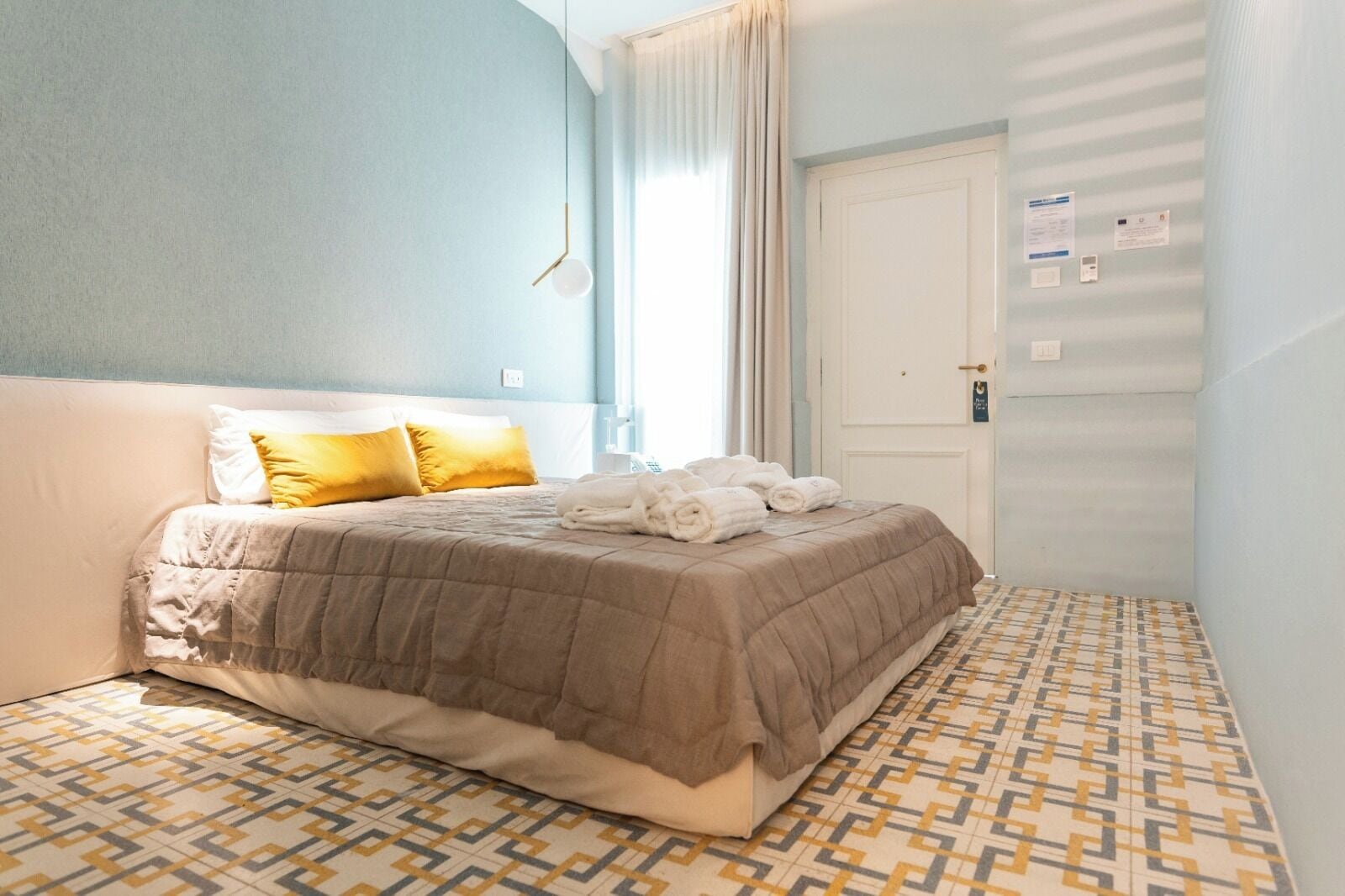 A hotel room with a bed, a patterned floor, a door with a lock, and a light switch.
