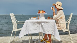Uma mesa posta sobre um deck de madeira, com toalha branca, um prato de frutas e uma pessoa bebendo em uma xícara.