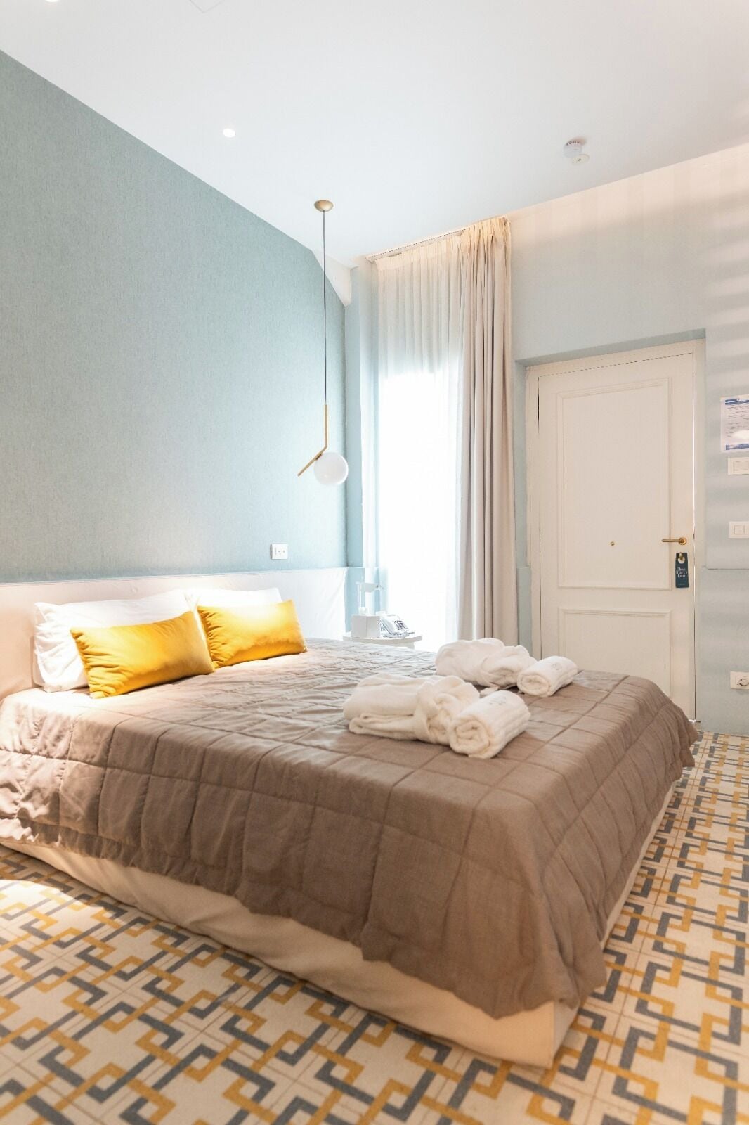 A hotel room with a large bed, a bedside table, a hanging light fixture, a window with curtains, and a patterned floor.
