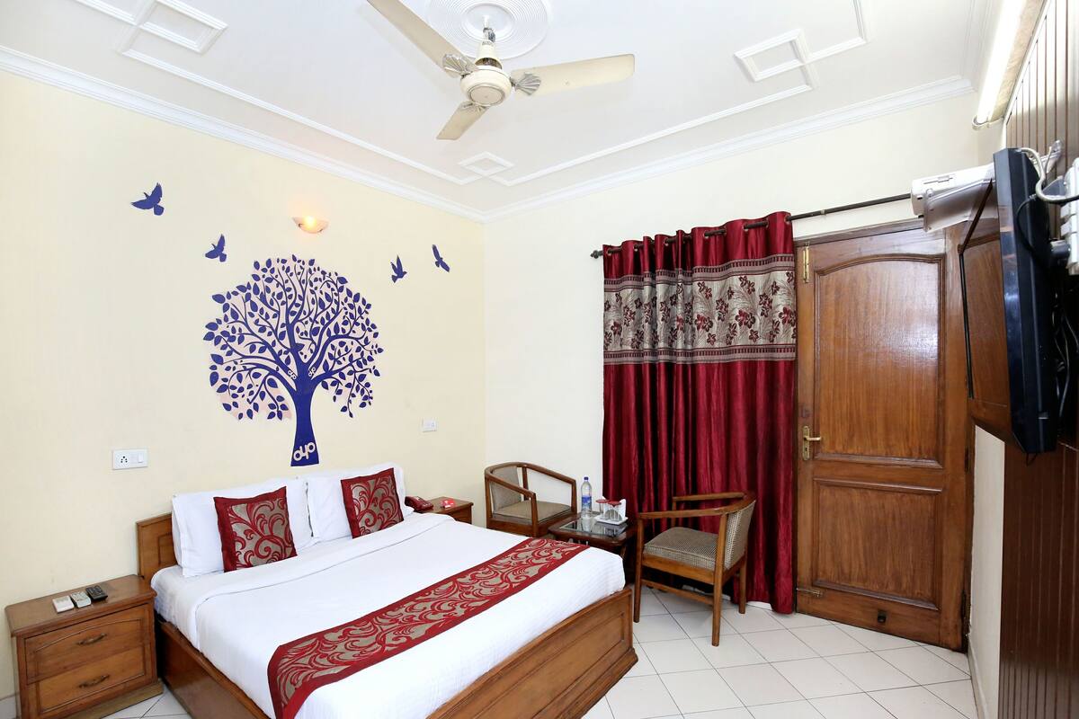 A bedroom with a bed, a wooden nightstand, a chair, a ceiling fan, a wall with a tree decal, and a door with curtains.
