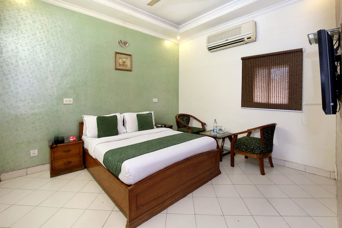 A hotel room with a wooden bed, a ceiling fan, an air conditioner, a small table with a chair, and a window with blinds.