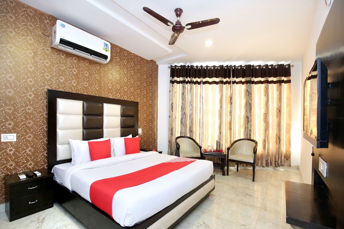 A hotel room with a large bed, a ceiling fan, an air conditioning unit, a TV, and a seating area with two chairs.
