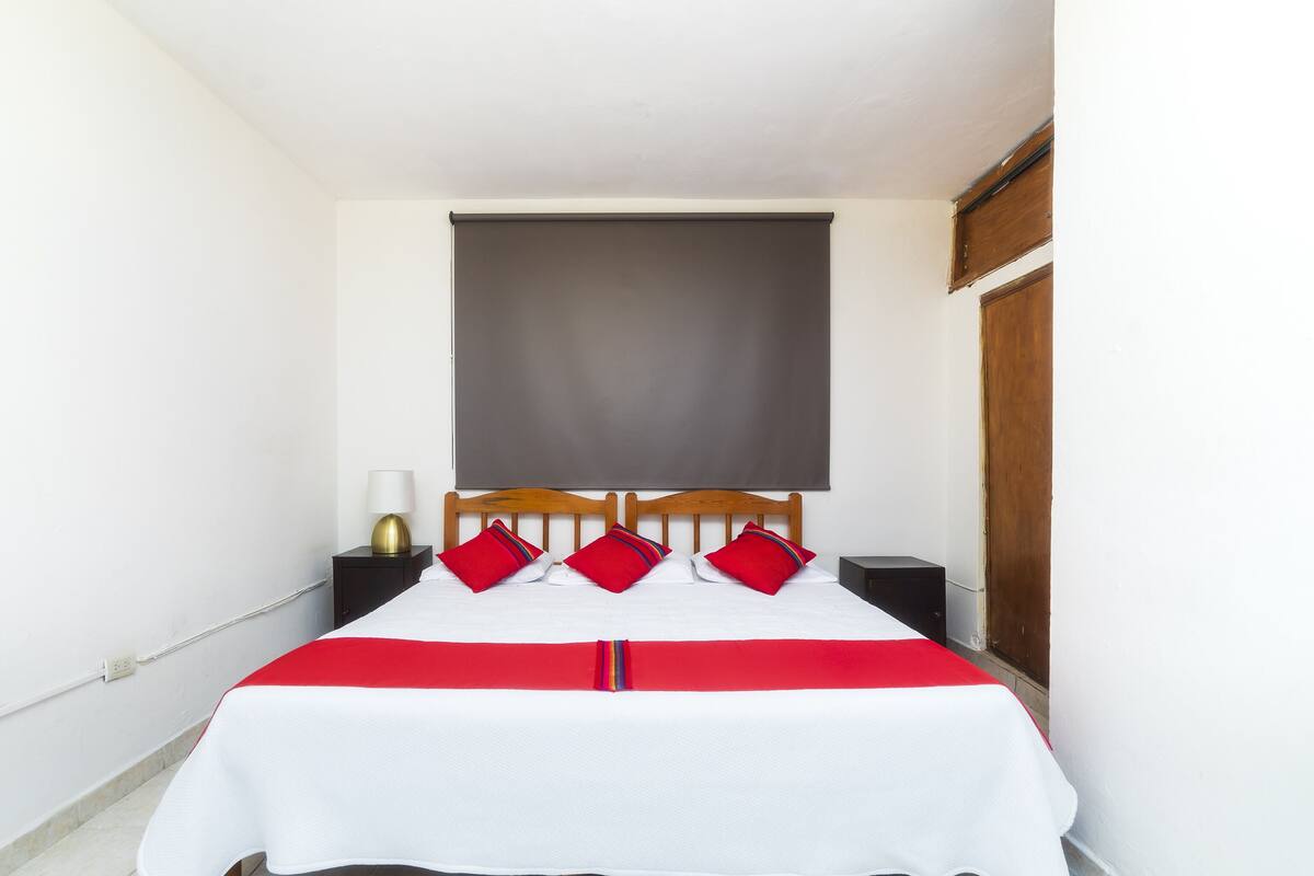 Ein ordentlich bezogenes Bett mit einer weiß-roten Tagesdecke, einem Holz-Kopfschild und zwei roten Kissen.