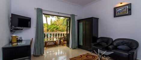 A living room with a TV, a sofa, a coffee table, and a view of a balcony with a railing and greenery.